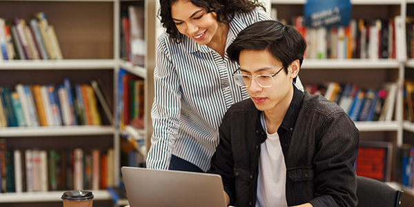 An academic advisor standing beside a student at a laptop in a library setting.